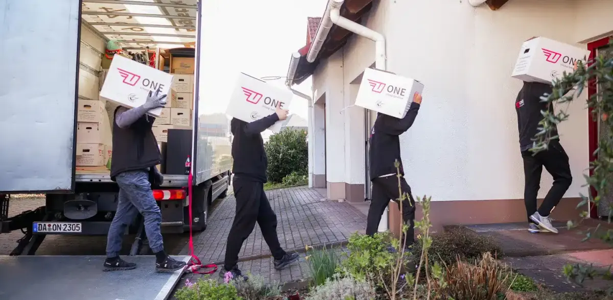 Vier Träger bringen ONE-Umzugskartons vom Möbelwagen nacheinander zur Haustür eines Einfamilienhauses.