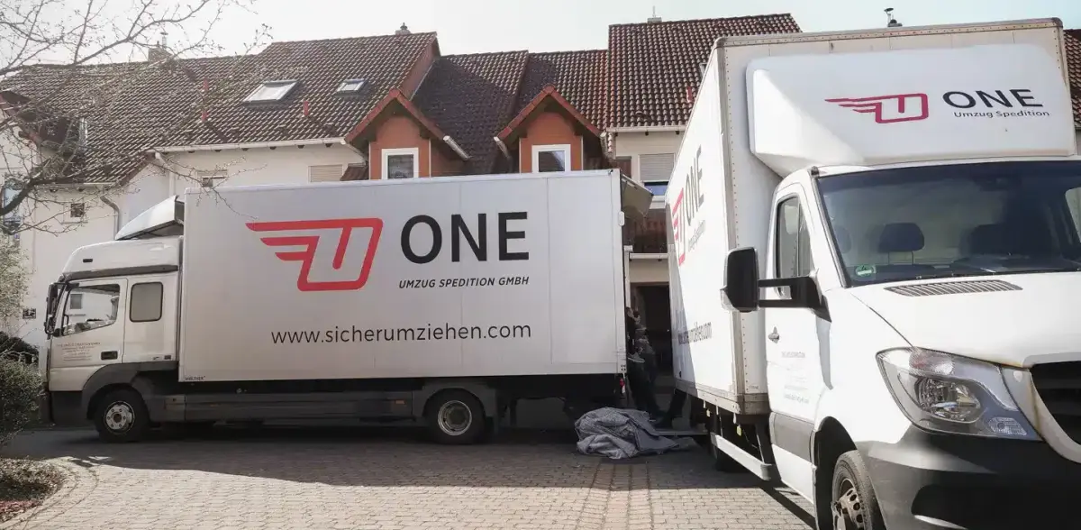 Zwei Möbelwagen der One Umzugsspedition stehen beladebereit in der Einfahrt eines Wohnhauses.