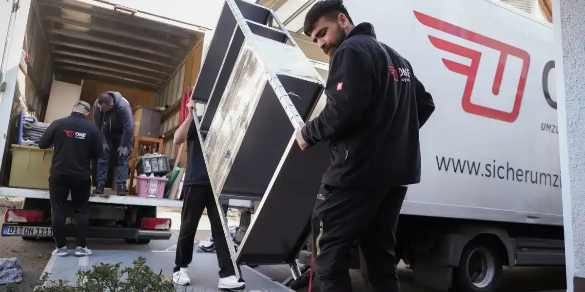 Zwei Mitarbeiter in ONE-Kleidung tragen ein großes dunkles Möbelstück über die Ladebordwand zum Möbelwagen, im Hintergrund verlädt das übrige Team weitere Möbel.