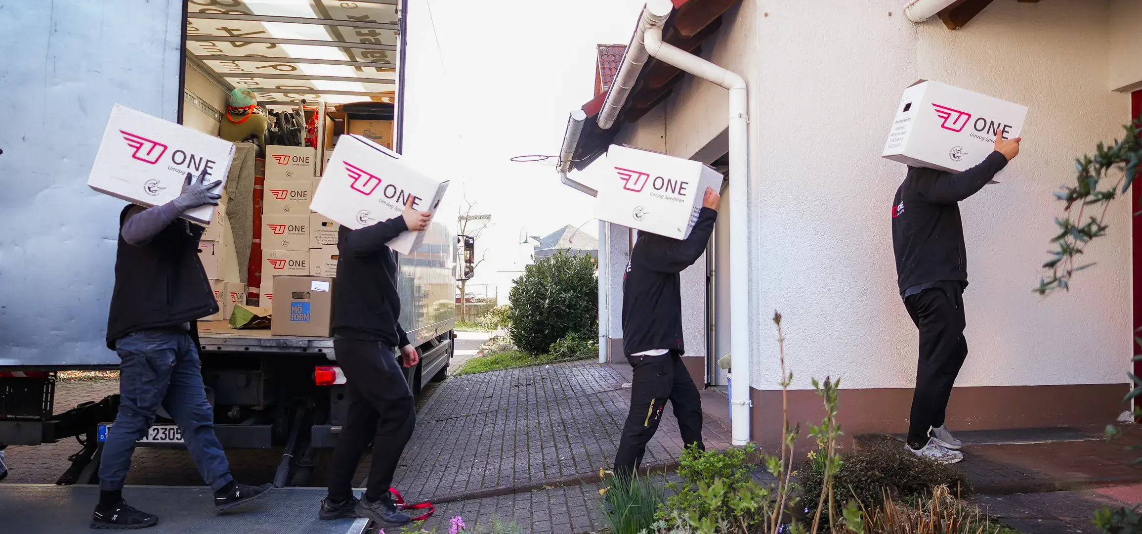 Vier Mitarbeitende der One Umzugsspedition tragen nacheinander weiße ONE-Umzugskartons vom Möbelwagen zu einem Wohnhaus.