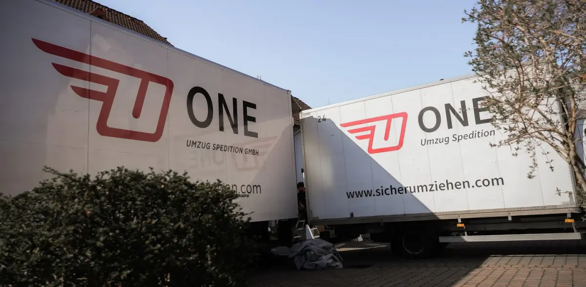 Zwei Möbelwagen mit ONE-Logo parken Seite an Seite, dazwischen arbeitet ein Träger an der Ladeklappe.