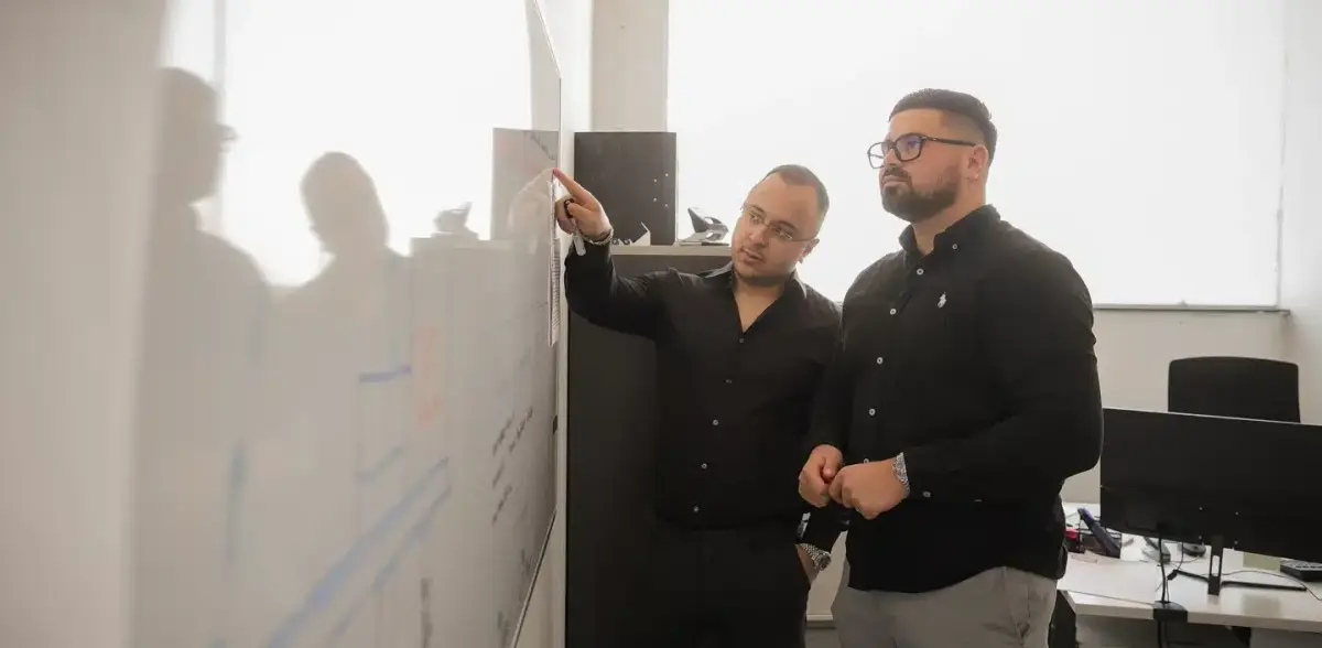 Zwei Mitarbeiter der One Umzugsspedition besprechen im Büro einen Ablaufplan an einer beschrifteten Planungstafel.