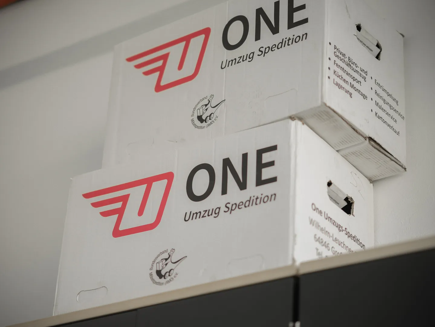 Zwei gestapelte weiße ONE-Umzugskartons mit dem roten Flügel-Logo; unter dem Logo ist das AMÖ-Verbandszeichen aufgedruckt.