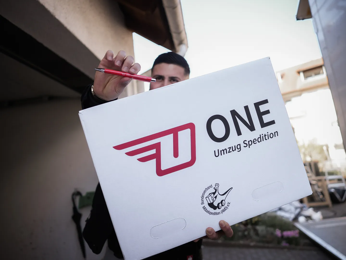 Mitarbeiter der One Umzugsspedition hält einen weißen Umzugskarton mit Firmenlogo und einen roten Stift in die Kamera