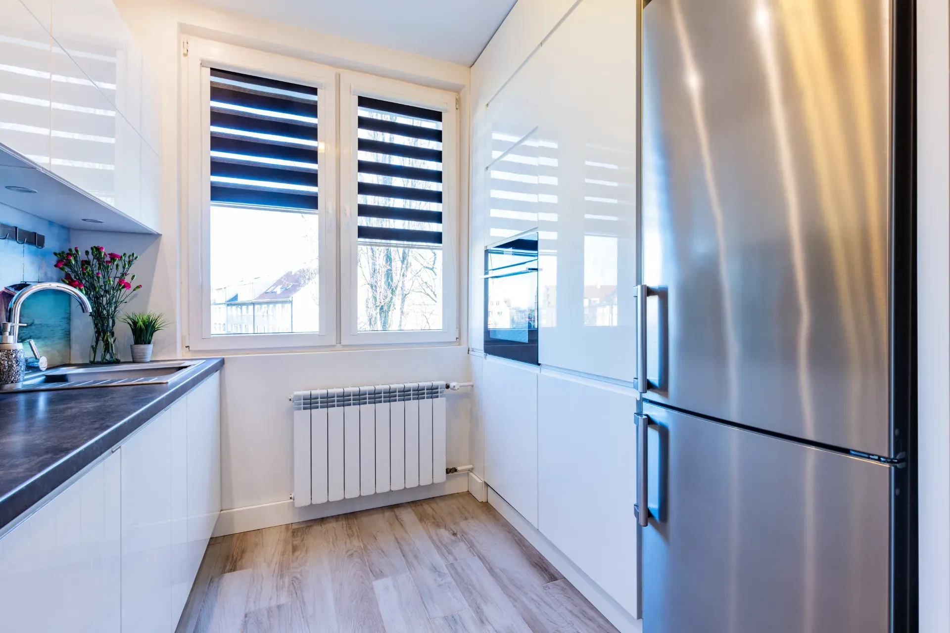 Schmale weiße Küche mit hohem Edelstahl-Kühlschrank neben Einbauschränken und Fenster.