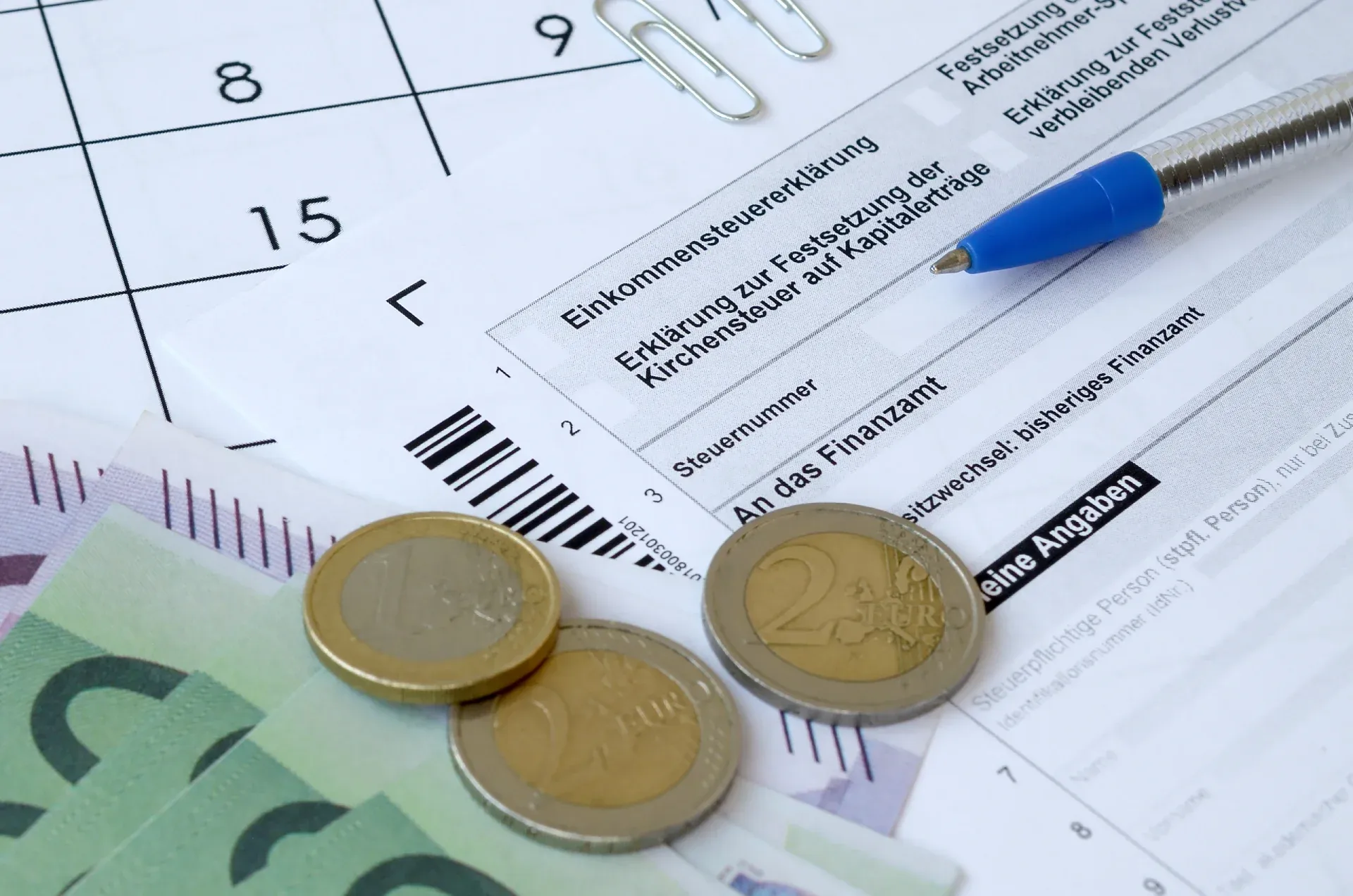 Formular der Einkommensteuererklärung mit Kugelschreiber, Euro-Münzen und Geldscheinen auf einem Kalenderblatt.