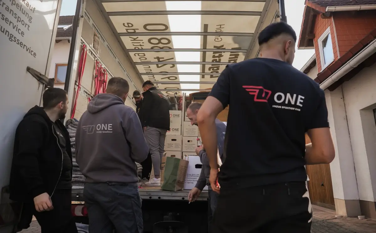 Mehrere Mitarbeitende in ONE-Shirts stapeln beschriftete Umzugskartons im offenen Möbelwagen, rote Spanngurte hängen an der Innenwand.