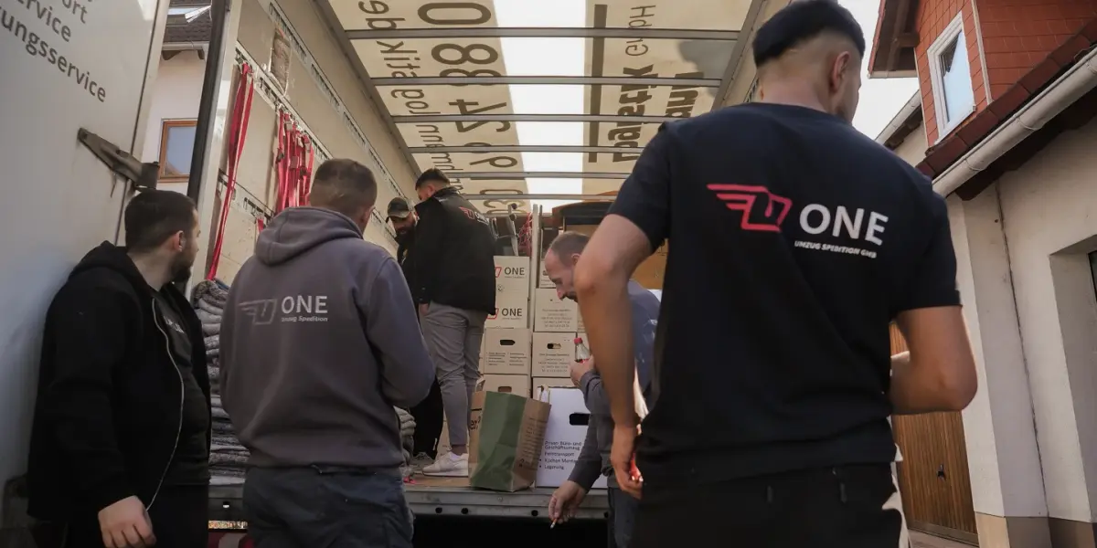 Mehrere Mitarbeitende in ONE-Shirts stapeln beschriftete Umzugskartons im offenen Möbelwagen, rote Spanngurte hängen an der Innenwand.