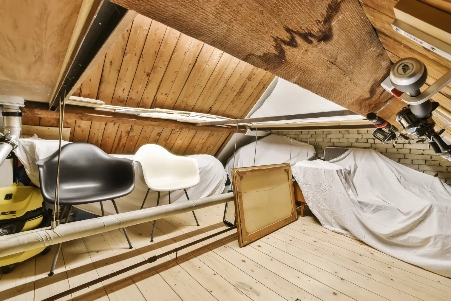 Dachboden mit Holzbalken: abgedeckte Möbel, zwei Stühle, ein Bilderrahmen und ein Sauger unter der Schräge.