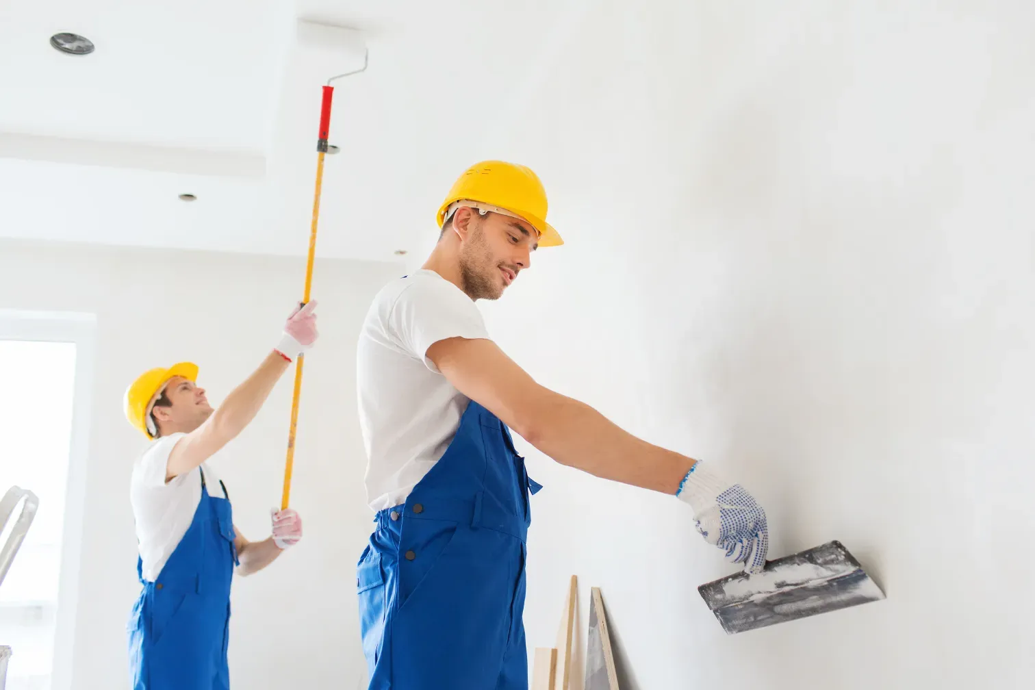 Zwei Handwerker in blauer Latzhose: Einer zieht Spachtelmasse auf die Wand, der andere streicht mit der Rolle die Decke.