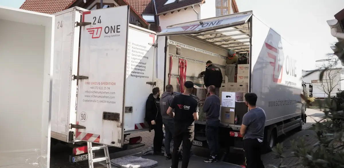 Umzugsteam in ONE-Kleidung belädt vor einem Einfamilienhaus zwei Möbelwagen mit Kartons.