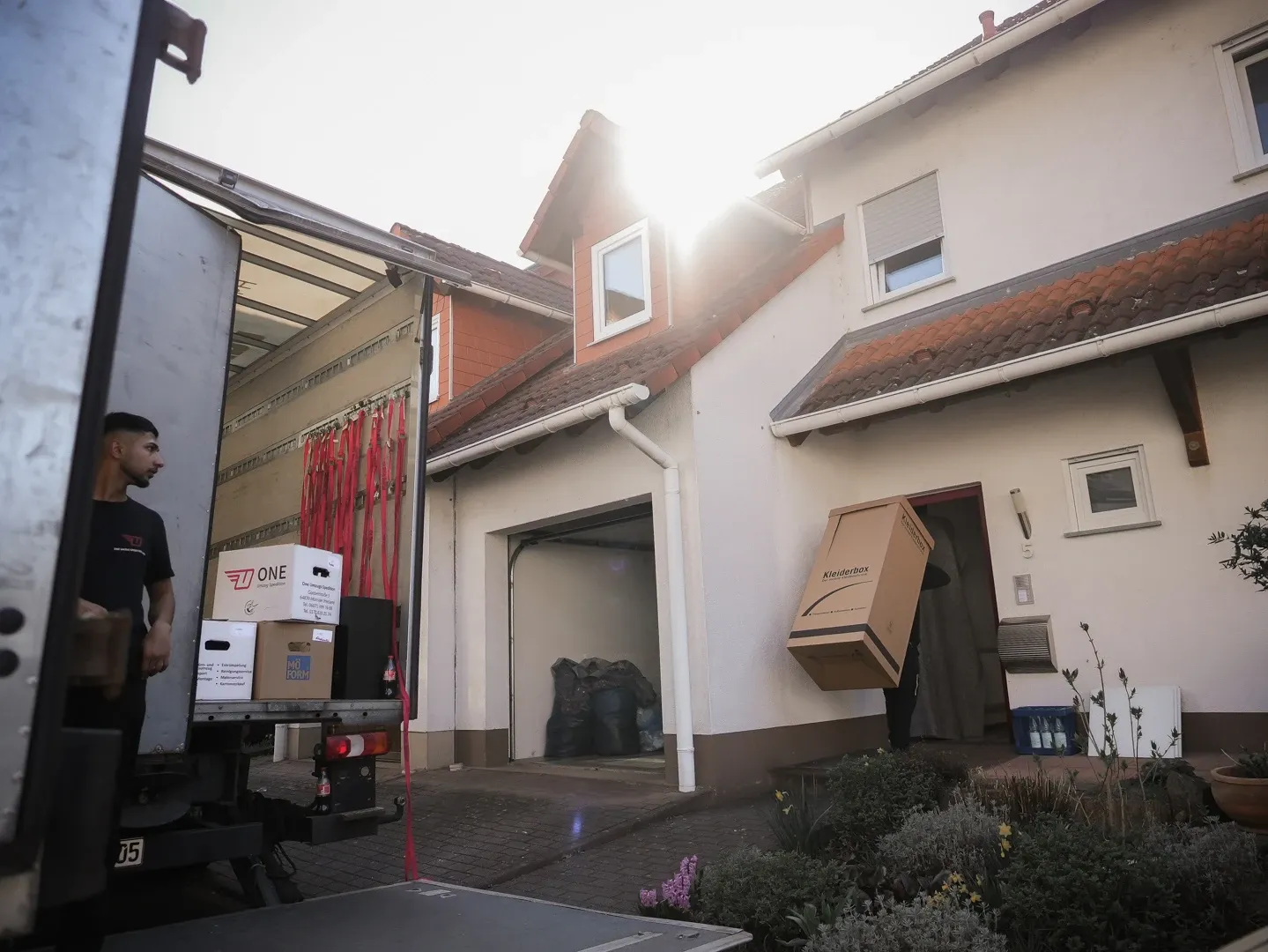 Mitarbeiter im geöffneten LKW am Zielort, ein Kollege trägt eine Kleiderbox in den Hauseingang.