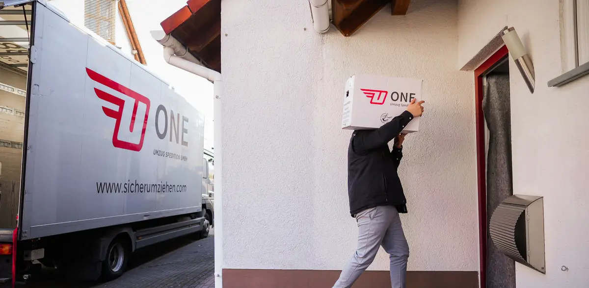Mitarbeiter trägt einen ONE-Umzugskarton vom Möbelwagen mit ONE-Logo zur geschützten Haustür eines Wohnhauses.