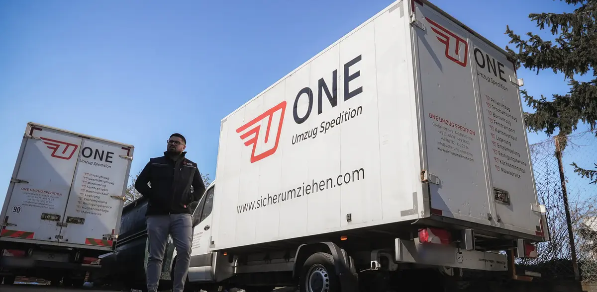 Mitarbeiter in ONE-Jacke steht vor zwei ONE-Möbelwagen mit Logo, Leistungsliste und Webadresse.