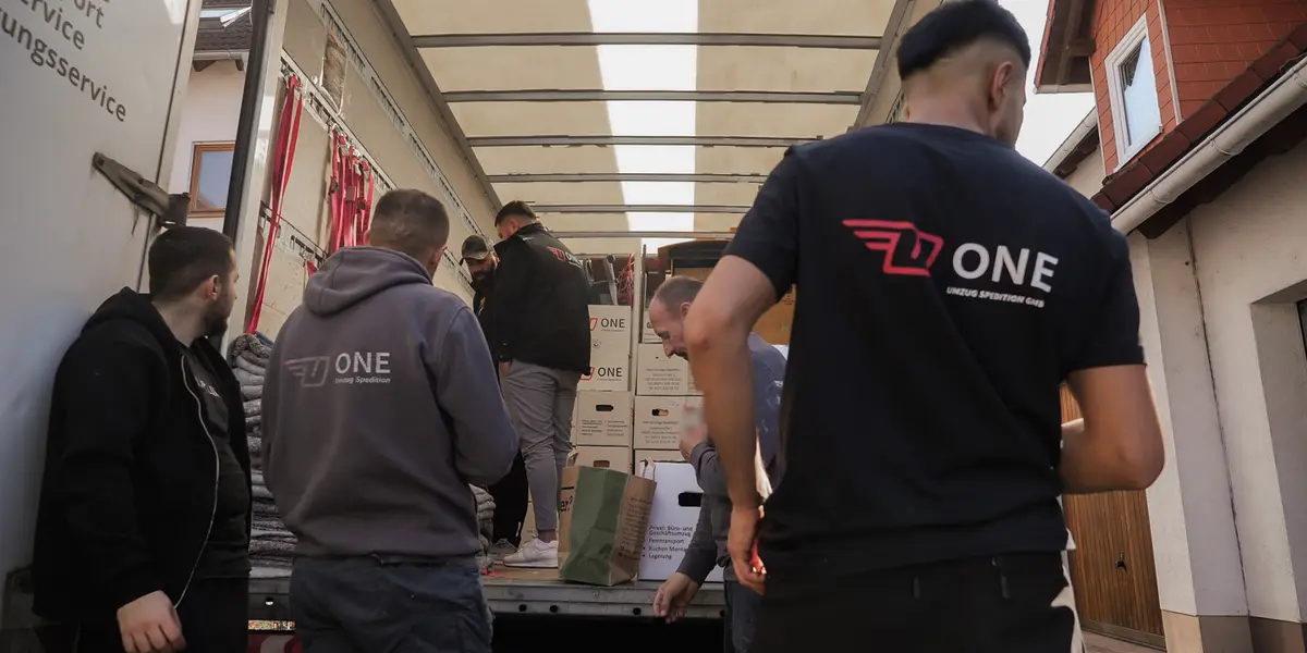 Mitarbeitende in ONE-Kleidung beladen einen Möbelwagen mit ONE-Umzugskartons, vorn ein Mitarbeiter mit großem ONE-Rückenaufdruck, an der Innenwand hängen rote Spanngurte.