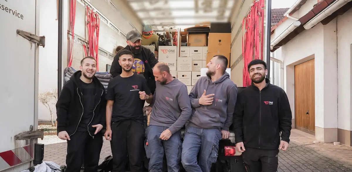 Sechs Mitarbeiter stehen lachend auf der Ladebordwand vor dem beladenen Möbelwagen, mehrere von ihnen in ONE-Kleidung.