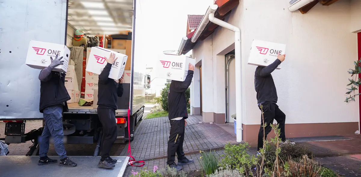 Vier Mitarbeiter tragen ONE-Umzugskartons in einer Reihe von der Ladebordwand des Möbelwagens zum Hauseingang.