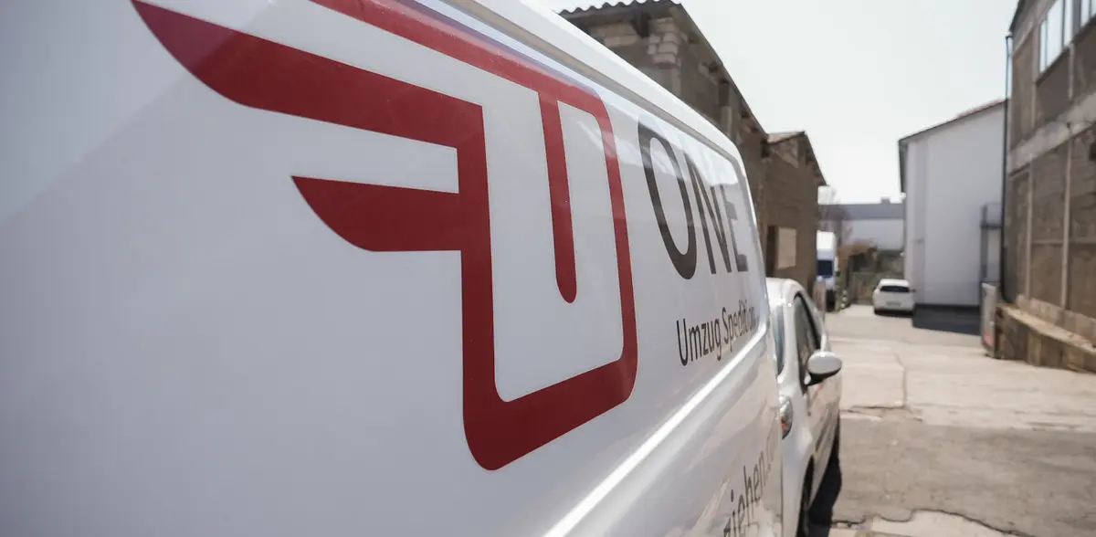 Seitenwand eines ONE-Transporters mit großem Flügel-Logo und dem Schriftzug ONE Umzug Spedition, dahinter ein Hof zwischen Gewerbegebäuden.