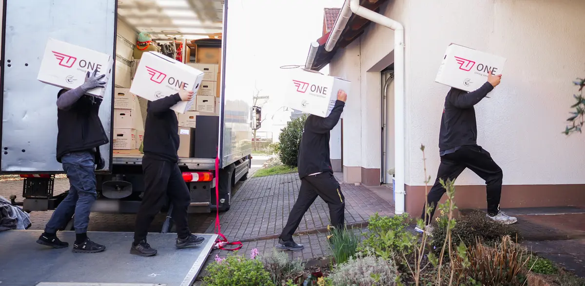 Vier Mitarbeiter tragen ONE-Umzugskartons hintereinander vom geöffneten Möbelwagen zur Haustür eines Einfamilienhauses.