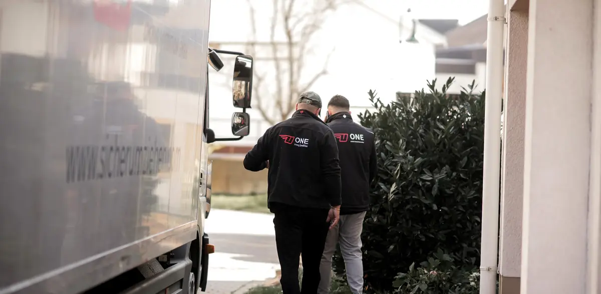 Zwei Mitarbeiter in ONE-Jacken gehen auf dem schmalen Weg zwischen Möbelwagen und Hauswand.