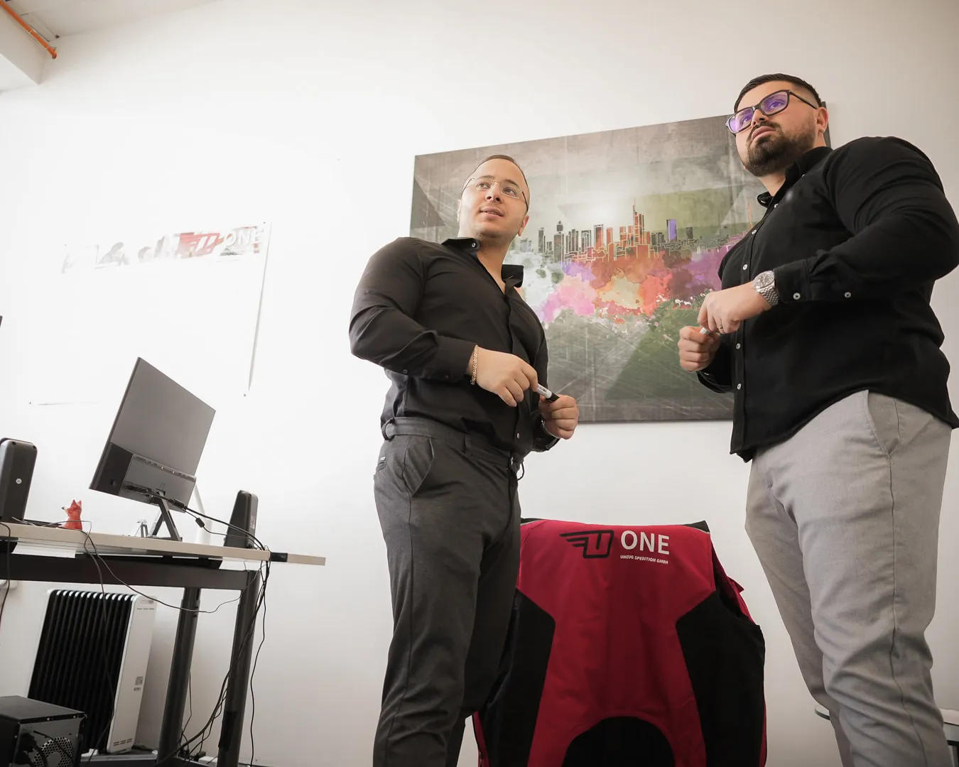 Zwei Mitarbeiter der One Umzugsspedition stehen im Büro vor einem Wandbild, links ein Schreibtisch mit Monitor, vorn ein Stuhl mit rot-schwarzer ONE-Jacke.