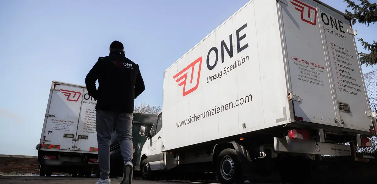 Mitarbeiter in ONE-Jacke geht auf zwei beschriftete Möbelwagen der One Umzugsspedition zu.