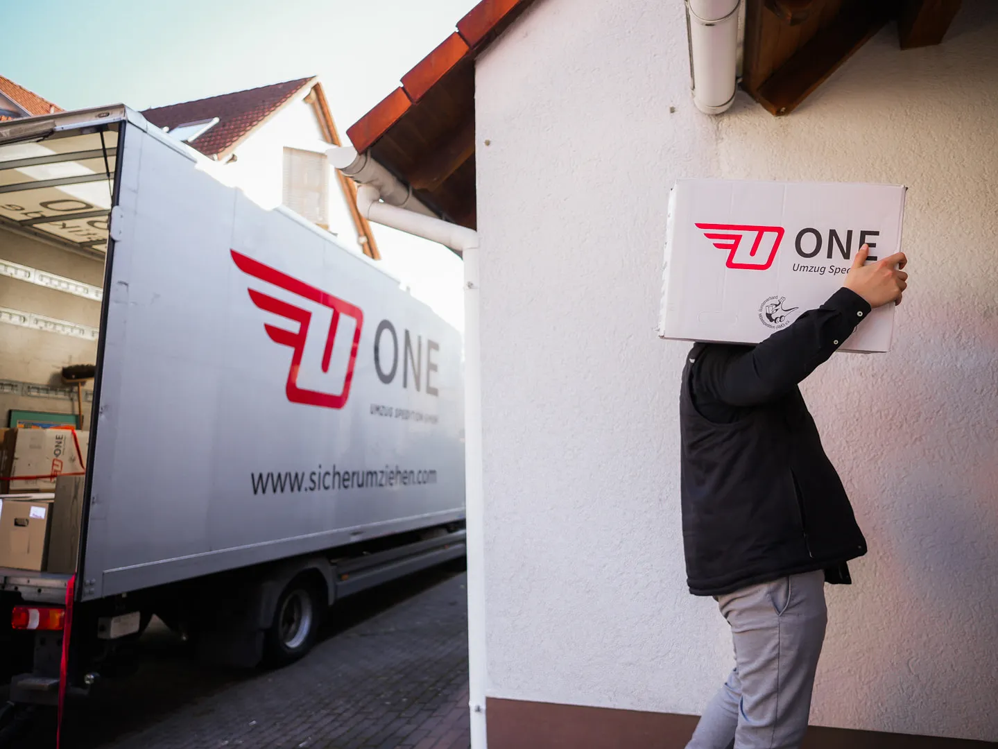 Mitarbeiter trägt einen ONE-Umzugskarton mit AMÖ-Zeichen an einer Hauswand entlang, links der Möbelwagen mit Logo und Internetadresse.