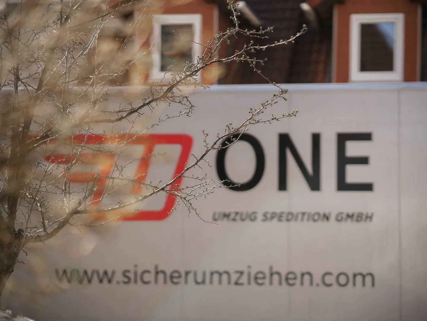 Möbelwagen der One Umzugsspedition mit Logo und Internetadresse vor einem Wohnhaus, im Vordergrund kahle Zweige mit Knospen.