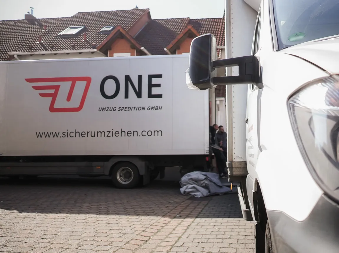Möbelwagen der One Umzugsspedition mit Logo und Internetadresse vor Wohnhäusern, rechts im Vordergrund die Front eines Transporters.