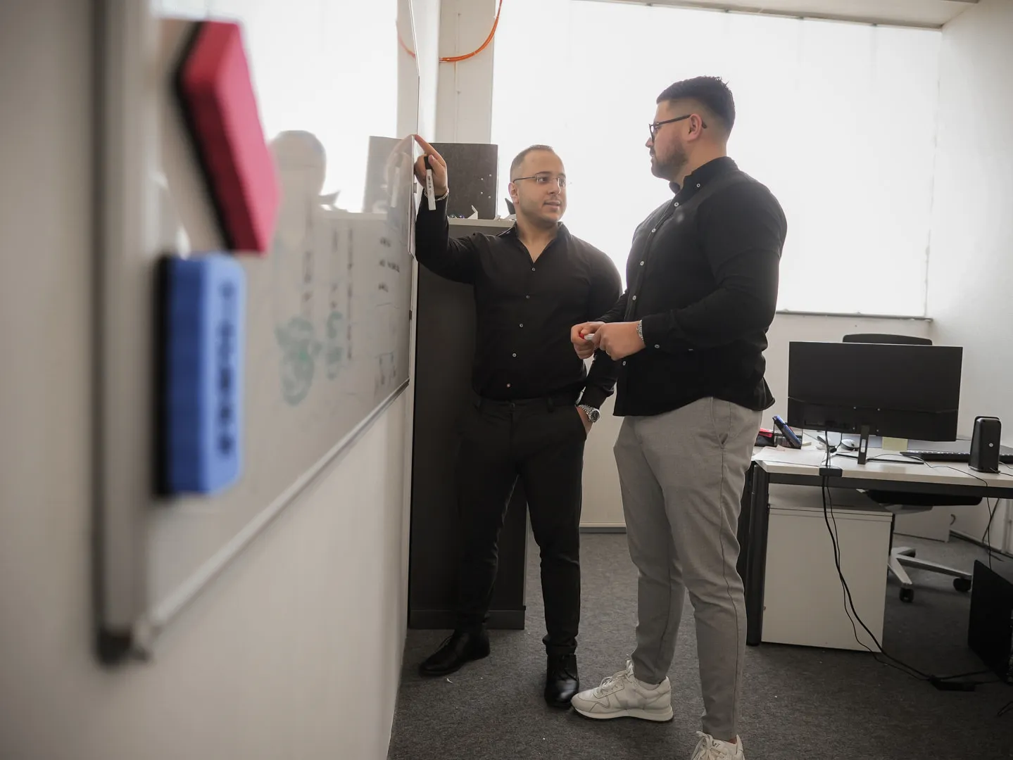 Zwei Mitarbeiter der One Umzugsspedition besprechen am Whiteboard im Büro den Ablauf, einer zeigt auf eine Notiz.