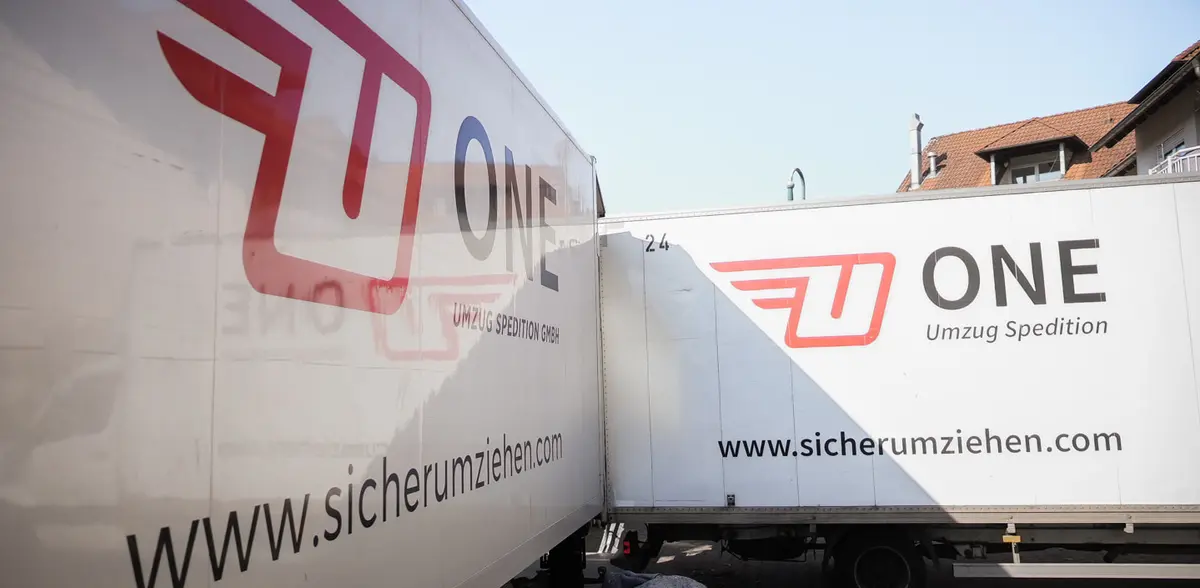 Zwei Möbelwagen der One Umzugsspedition stehen im rechten Winkel zueinander, beide mit großem Logo und Internetadresse.