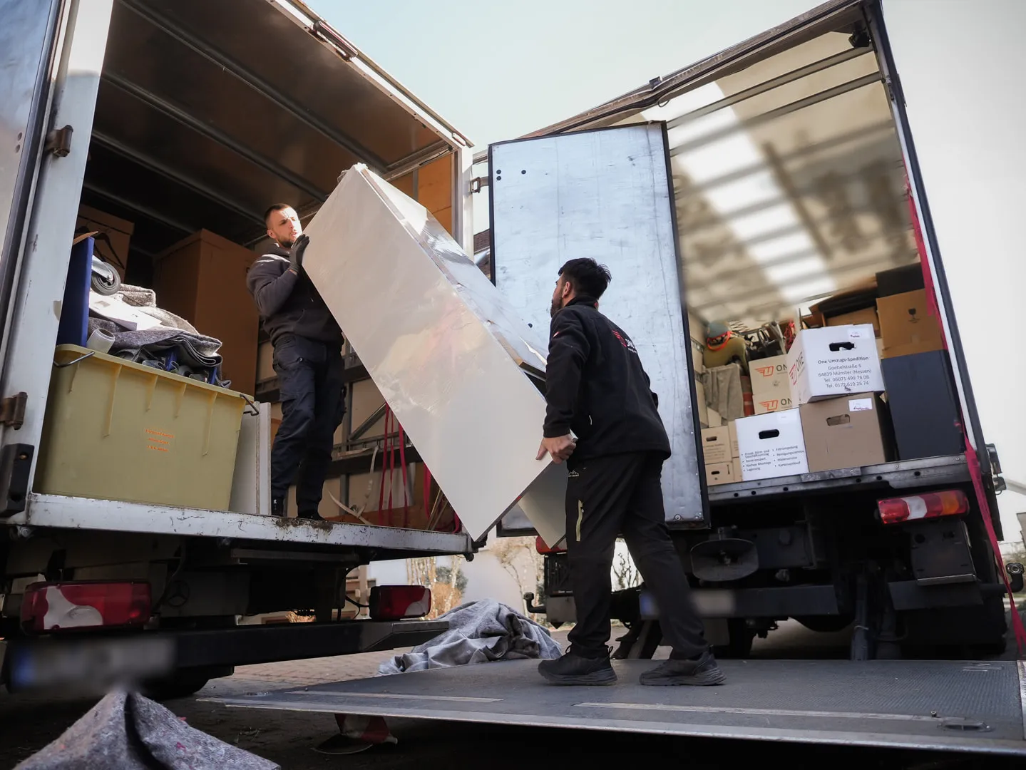 Zwei Mitarbeiter der One Umzugsspedition heben gemeinsam eine große Möbelplatte zwischen zwei geöffneten LKW-Hecks in den Laderaum.