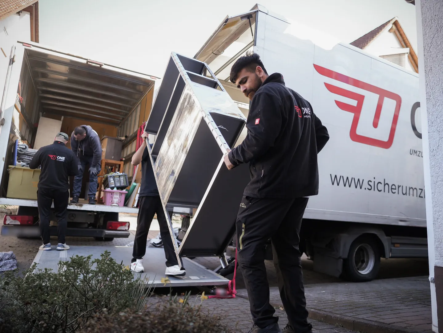 Zwei Mitarbeiter der One Umzugsspedition tragen ein Metallregal über die Ladebordwand zum Möbelwagen.