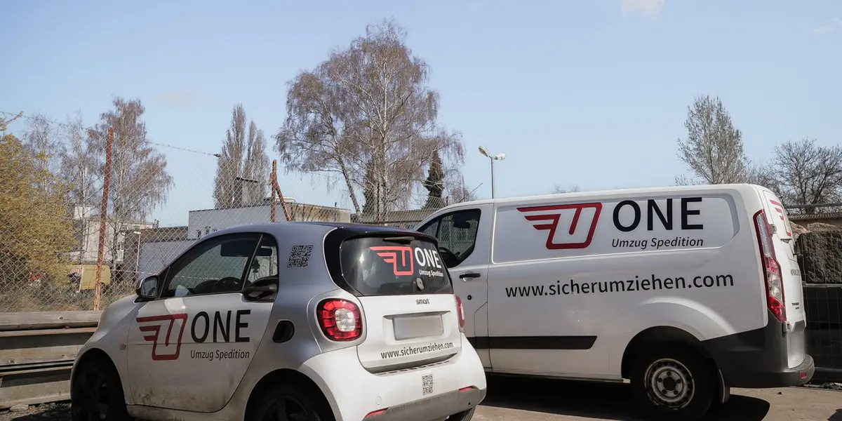 Ein Kleinwagen und ein Transporter mit ONE-Logo und Webadresse stehen auf dem Betriebshof der One Umzugsspedition.
