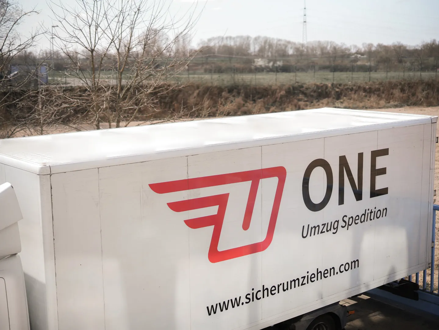 Blick von oben auf den Kofferaufbau eines Möbelwagens mit großem ONE-Logo und Webadresse, dahinter Felder und kahle Bäume.