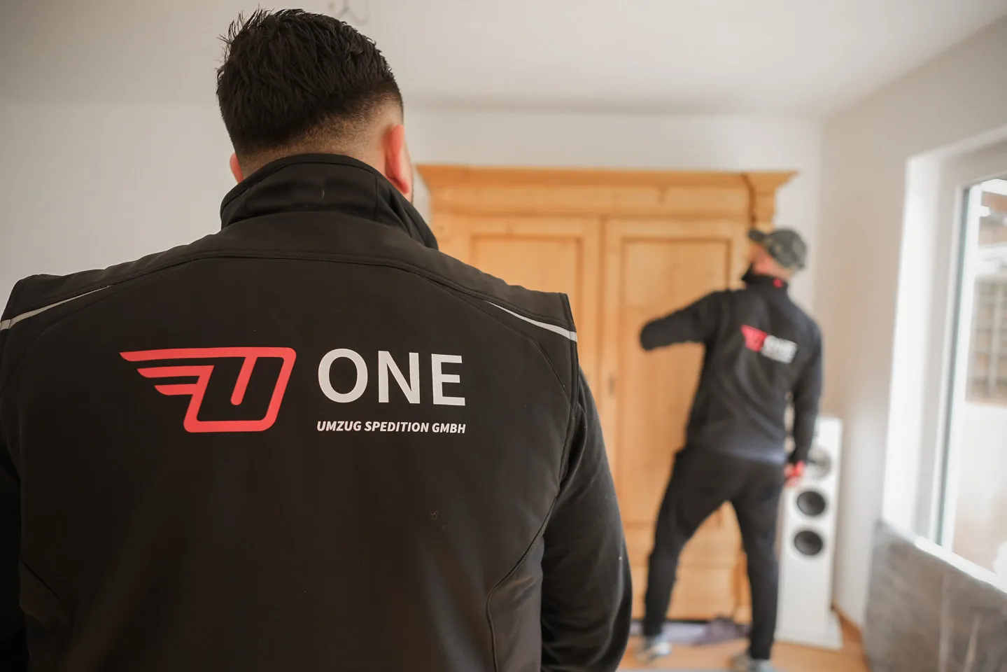 Mitarbeiter mit ONE-Logo auf dem Rücken, dahinter arbeitet ein Kollege in einer Wohnung an einem Holzschrank.