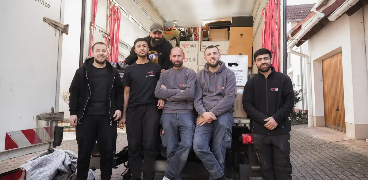 Sechs Mitarbeiter der One Umzugsspedition stehen vor der geöffneten Ladebordwand ihres beladenen Umzugswagens; an der Innenwand hängen rote Spanngurte, dahinter stehen gestapelte ONE-Umzugskartons.