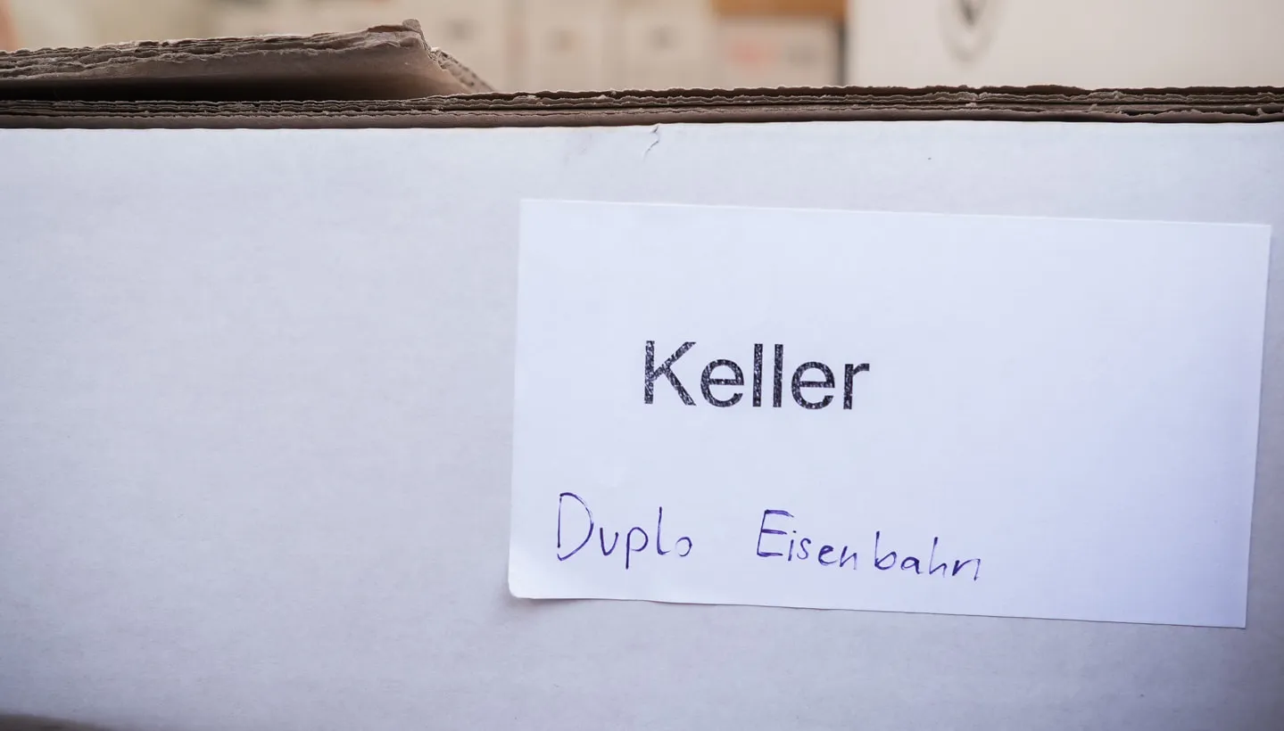 Umzugskarton mit aufgeklebtem Etikett „Keller“ und handschriftlicher Inhaltsangabe, am oberen Rand unscharf weitere Kartons