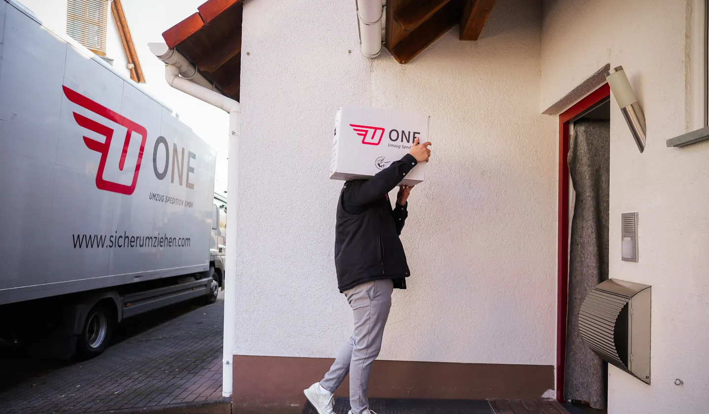 Mitarbeiter von ONE geht mit einem Umzugskarton mit Firmenlogo auf die mit einer Möbeldecke geschützte Haustür zu, rechts Klingel und Briefkasten, links der ONE-Lkw
