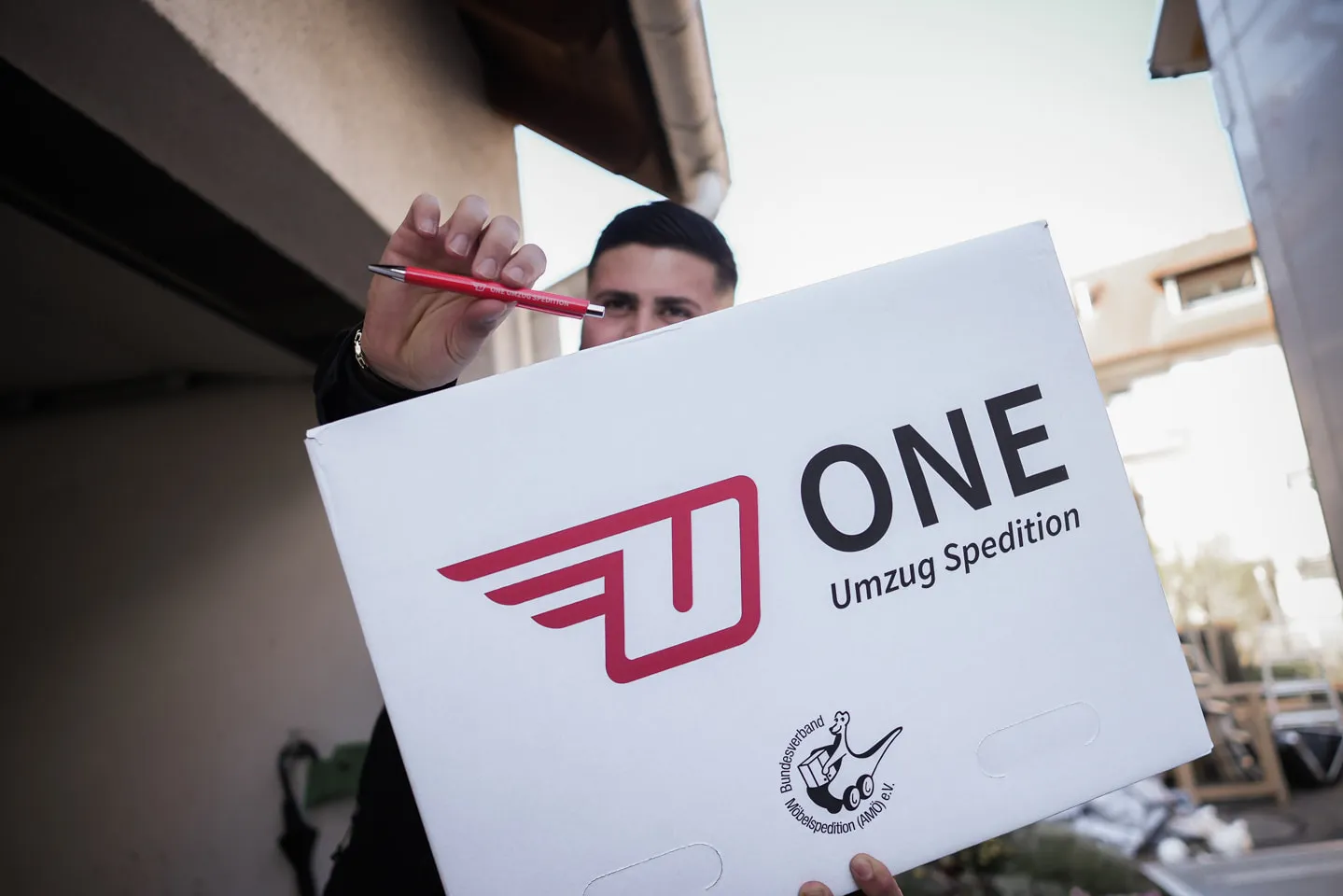 Mitarbeiter der One Umzugsspedition hält einen weißen Umzugskarton mit Firmenlogo und einen roten Stift in die Kamera