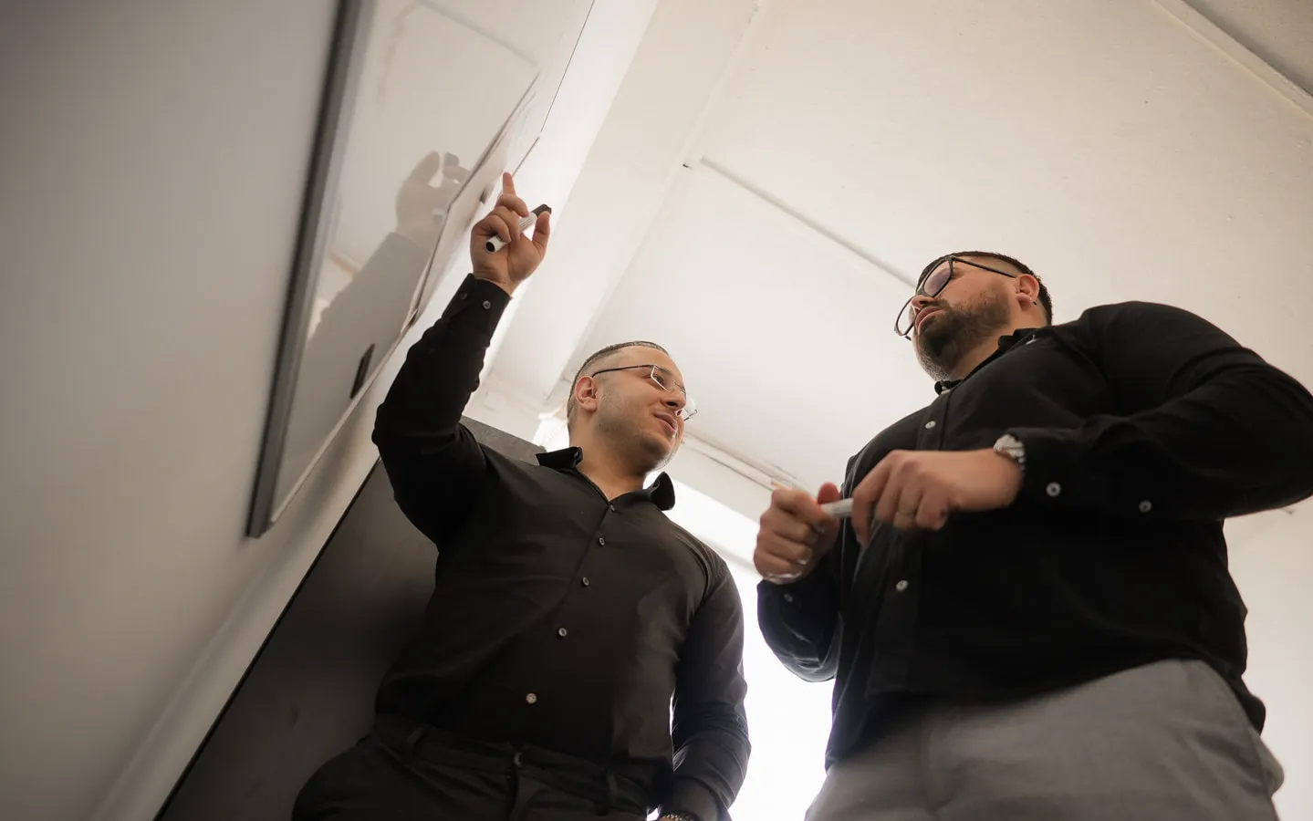 Zwei Mitarbeiter in schwarzen Hemden am Whiteboard im Büro: Einer zeigt mit dem Marker auf das Board, der andere hält einen Marker in den Händen