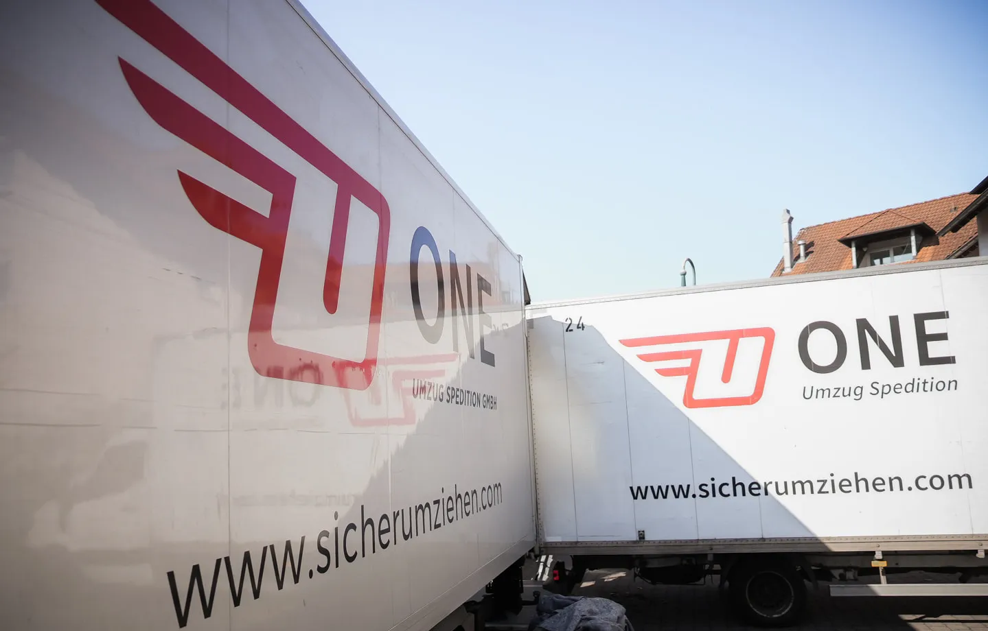 Zwei ONE-Lkw mit Firmenlogo und der Webadresse www.sicherumziehen.com, im Winkel zueinander geparkt
