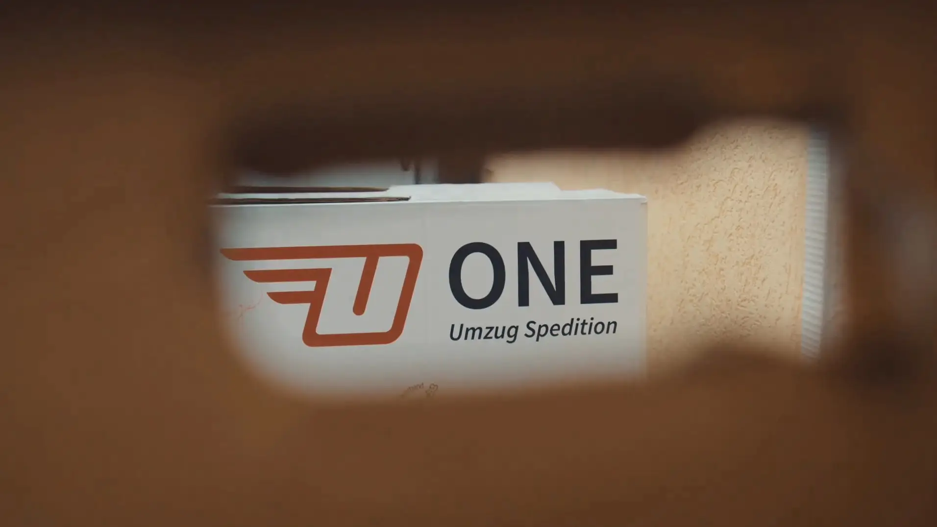 Blick durch die Grifföffnung eines Umzugskartons auf einen weißen ONE-Karton mit rotem Logo.
