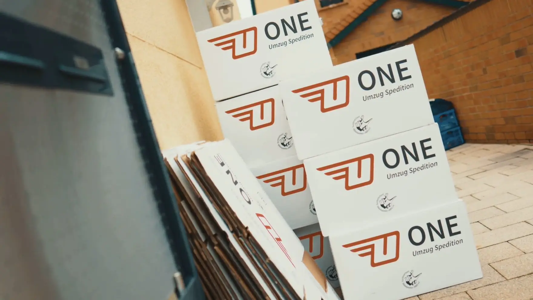 Gestapelte weiße ONE-Umzugskartons mit rotem Logo und AMÖ-Zeichen vor einem Hauseingang, daneben ein Stapel flach gefalteter Kartons.