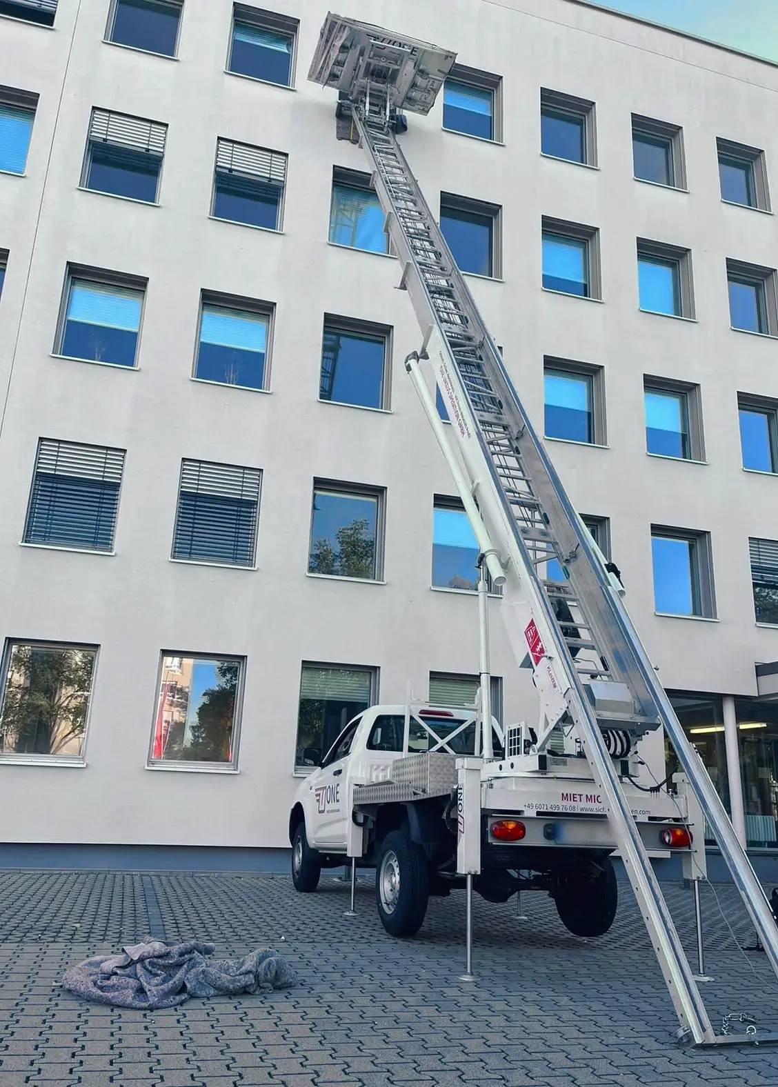 Weißer Pritschenwagen mit ausgefahrenen Stützen vor einem weißen Bürogebäude, die Leiter des Möbellifts ist bis zu den oberen Fenstern ausgefahren.