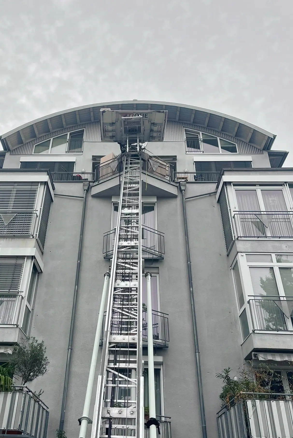 Blick von unten entlang der Leiter des Möbellifts zur Dachterrasse eines Wohnhauses mit Rundbogendach, dahinter bedeckter Himmel.