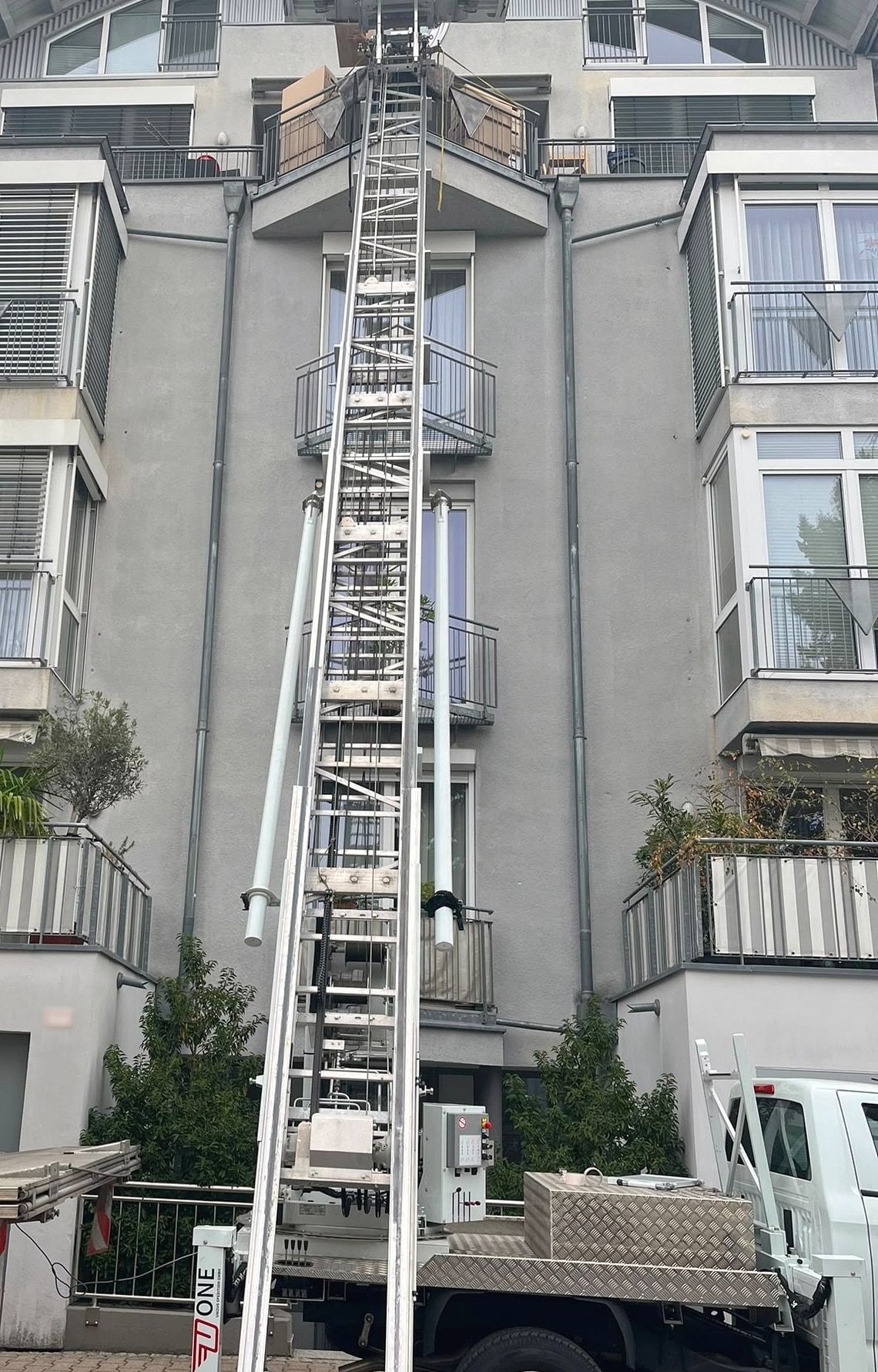 Möbellift der One Umzugsspedition reicht vom Pritschenwagen bis zur Dachterrasse eines Wohnhauses, auf der Plattform stehen Umzugskartons.