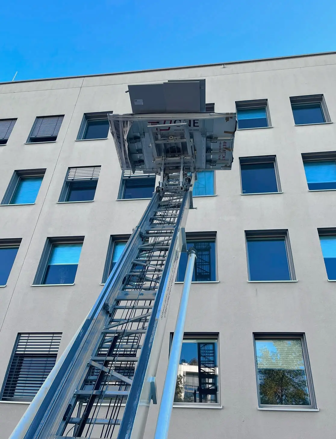 Plattform des Möbellifts mit einem hohen weißen Schrank frei vor blauem Himmel, links die Leiter, rechts die Fassade des Gebäudes.