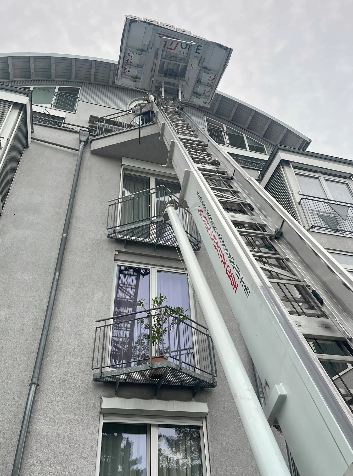 Plattform des Möbellifts mit Umzugskartons oben am Wohnhaus, am Hydraulikzylinder die Firmenaufschrift, rechts Balkone mit Pflanzen.
