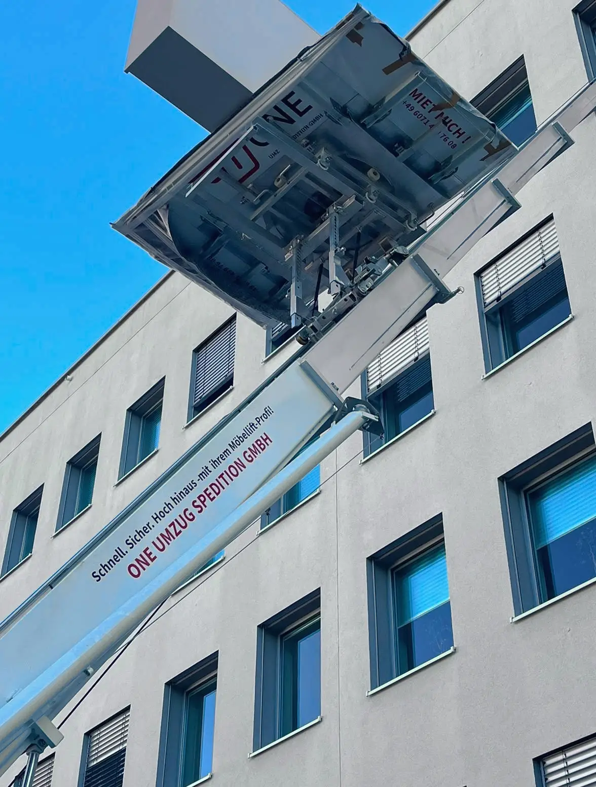 Nahaufnahme der Plattform des Möbellifts mit der Aufschrift „MIET MICH!“, darüber der Ausleger mit Firmenschriftzug vor einer hellen Fassade.
